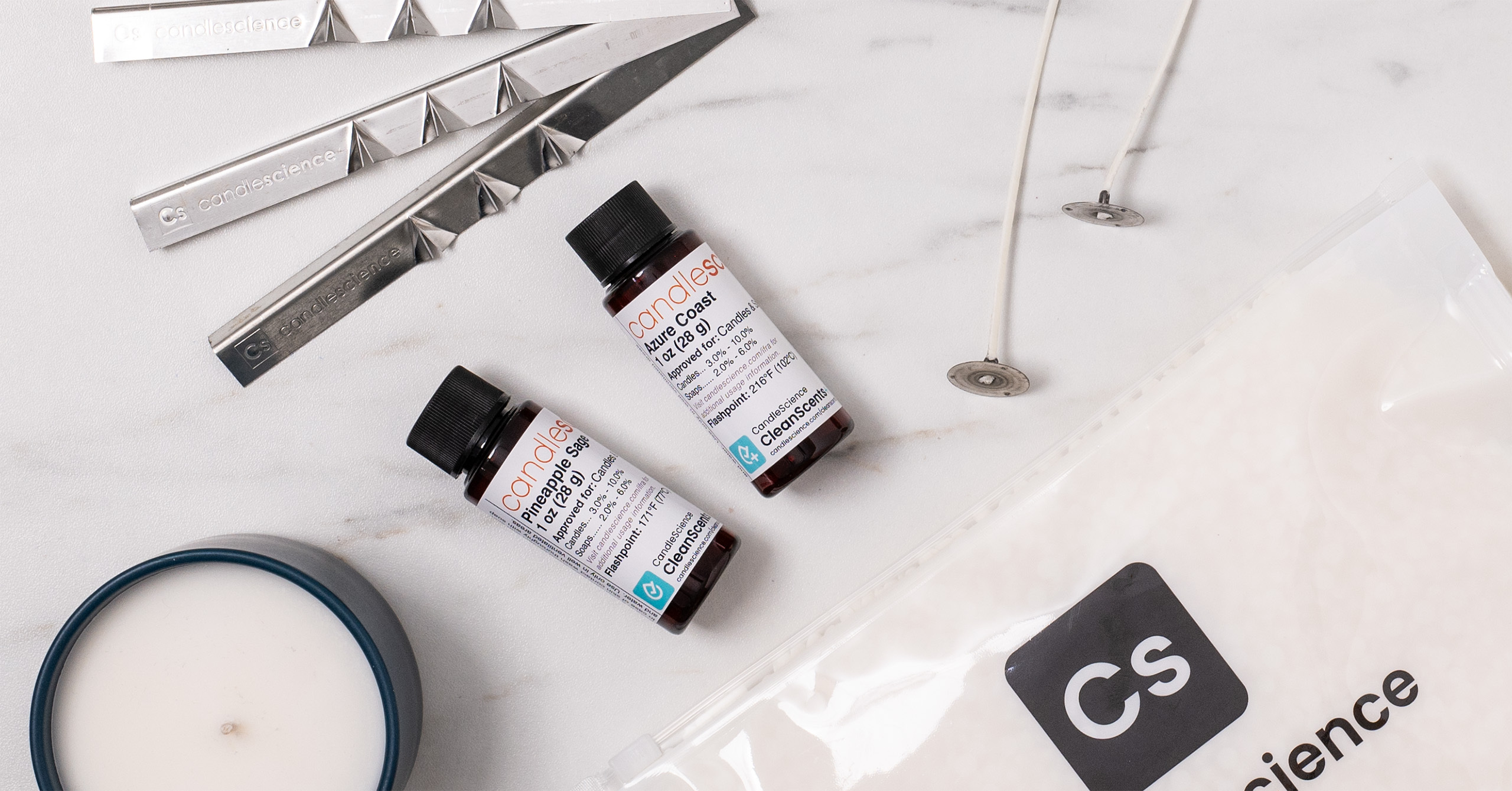 Two fragrance oil bottles, each showing a different CleanScents+ and CleanScents logo, on a white marble countertop with wicks, wax bags, and wick setters. 